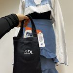 Črna vreča z belim napisom SDL. Vrečo drži roka. V ozadju je modno in modro oblečena lutka.