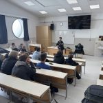Dijaki sedijo v predavalnici in poslušajo predavanje policista. Na sliki je tudi tabla, omara, računalnik.