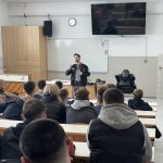 Dijaki sedijo v predavalnici in poslušajo predavanje policista. Na sliki je tudi tabla, omara in računalnik.