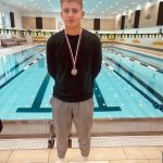 Dijak stoji pred bazenom in okoli vratu ima medaljo.