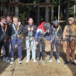 Dijaki stojijo v vrsti, so v naravi, oblečeni so v plezalna oblačila, saj gredo v adrenalinski park.