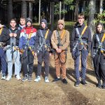 Dijaki stojijo v vrsti, so v naravi, oblečeni so v plezalna oblačila, saj gredo v adrenalinski park.