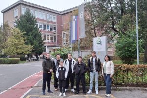 Dijaki programa okoljevarstveni tehnik in profesorja spremljevalca stojijo pred vhodom v Srednjo gradbeno, geodetsko, okoljevarstveno šolo in strokovno gimnazijo Ljubljana. Za dijaki je izobešena slovenska zastava in zastava SGGOŠ.StG LJ. Na levi in desni strani za dijaki so zelena listnata in iglasta
