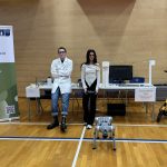 Dijaka stojita pred mizo, na kateri je kemijski vrt, elektro labirint, na tleh je robotski pes. Desno od mize je električni skiro.