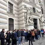 Dijaki stojijo pred stavbo in poslušajo profesorico, ki razlaga.