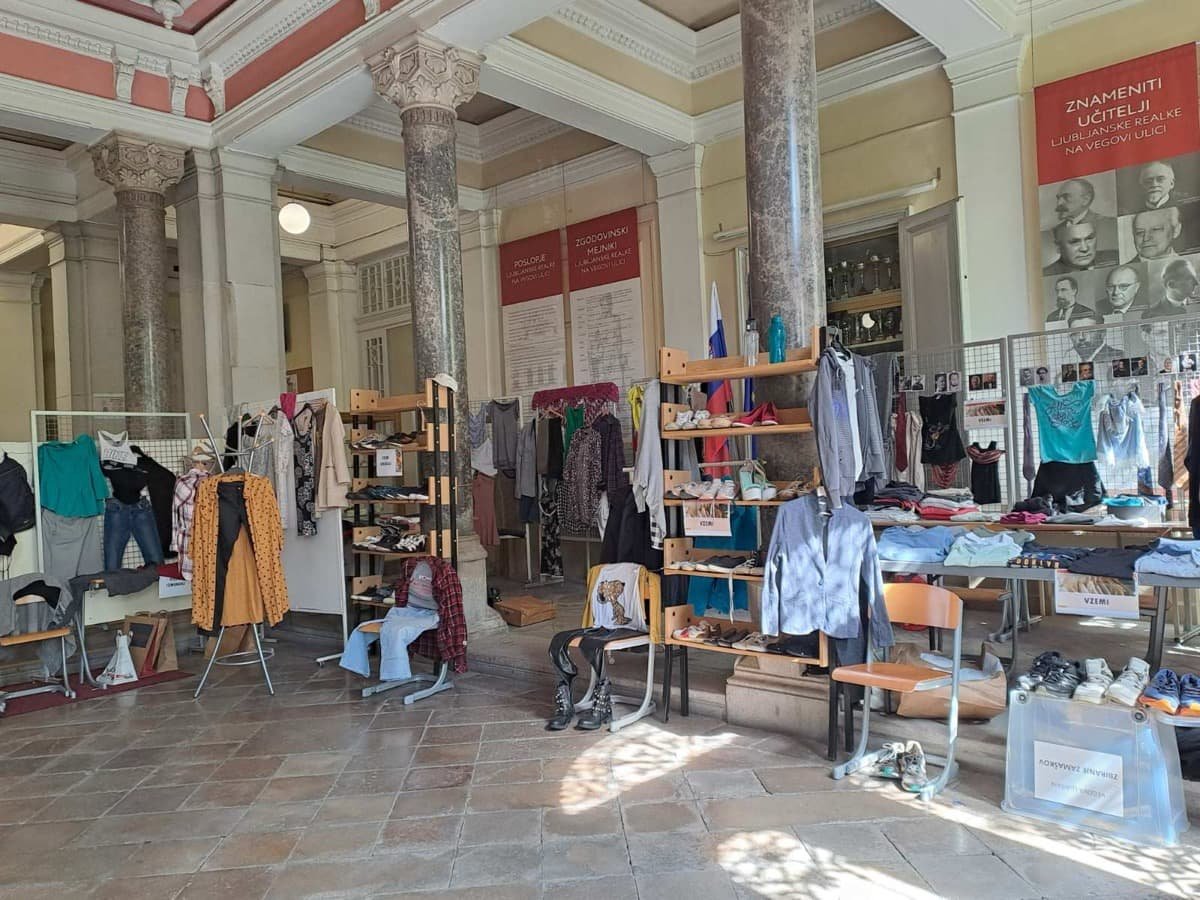 Zbirni center trajnostne izmenjevalnice v šolski avli, kjer so na mizah, stolih in panojih razstavljena oblačila in obutev. V ozadju so plakati znanih Vegovcev, zastavi in vitrina s pokali.